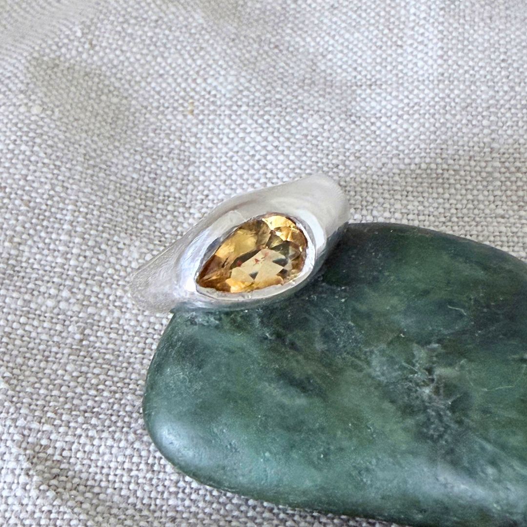 Close-up of Kōwhai Signet Ring showing pear-cut natural citrine and organic silver setting beside pounamu