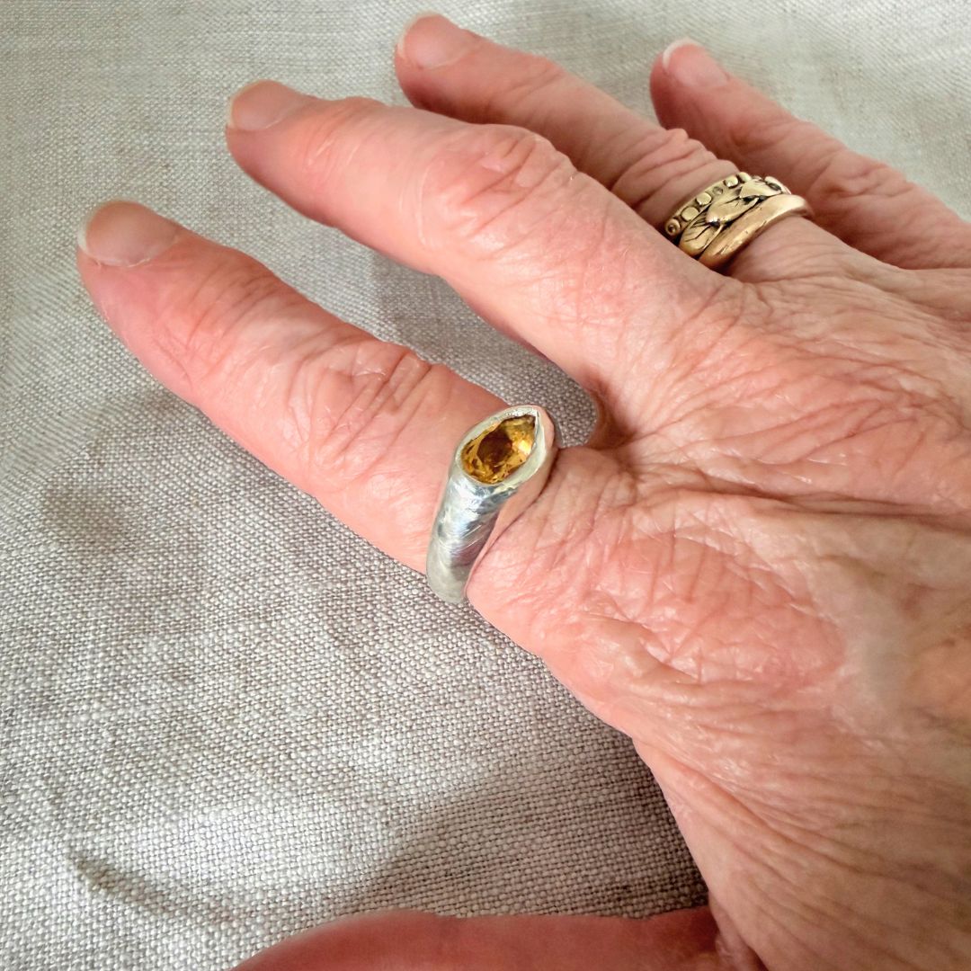 A close-up of a hand wearing a unique silver ring set with a faceted Citrine. The hand is resting on a textured beige fabric background, with other textured gold rings visible.