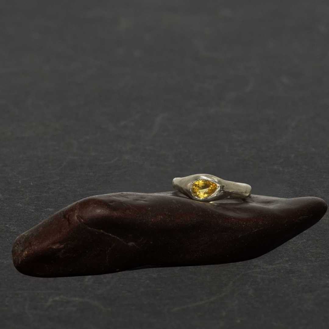 The Kowhai organic sterling silver signet ring featuring a glowing pear-shaped yellow citrine gemstone, balanced on a smooth dark-red stone against a dark grey background.