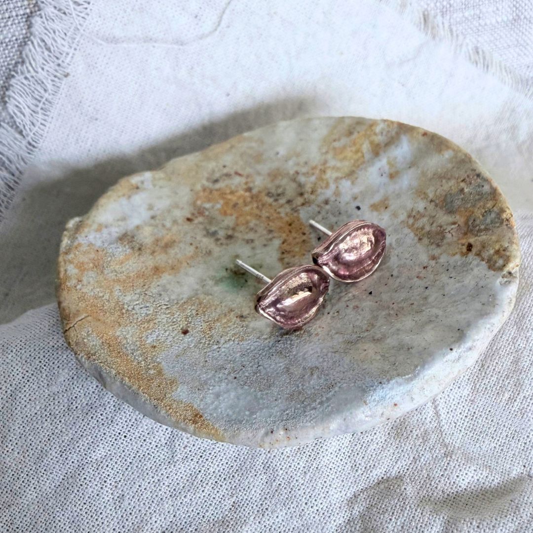 Bronze Kōwhai Pod stud earrings with sterling silver posts displayed on a pale stone dish and linen background.
