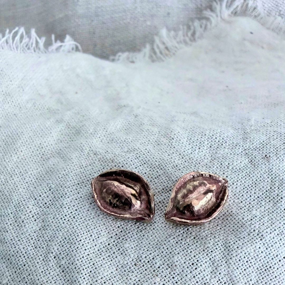 Pair of bronze Kōwhai Pod stud earrings resting on natural linen, showing the hand-finished pod texture.