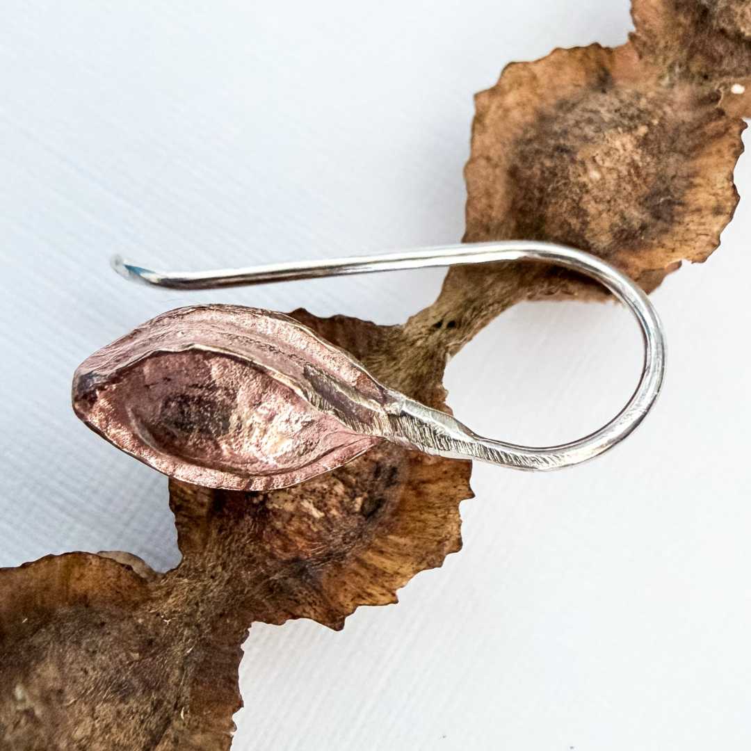 A handcrafted Sterling Silver and Bronze kowhai pod drop earring resting directly on a dried, textured natural kowhai seed pod to show the design inspiration.