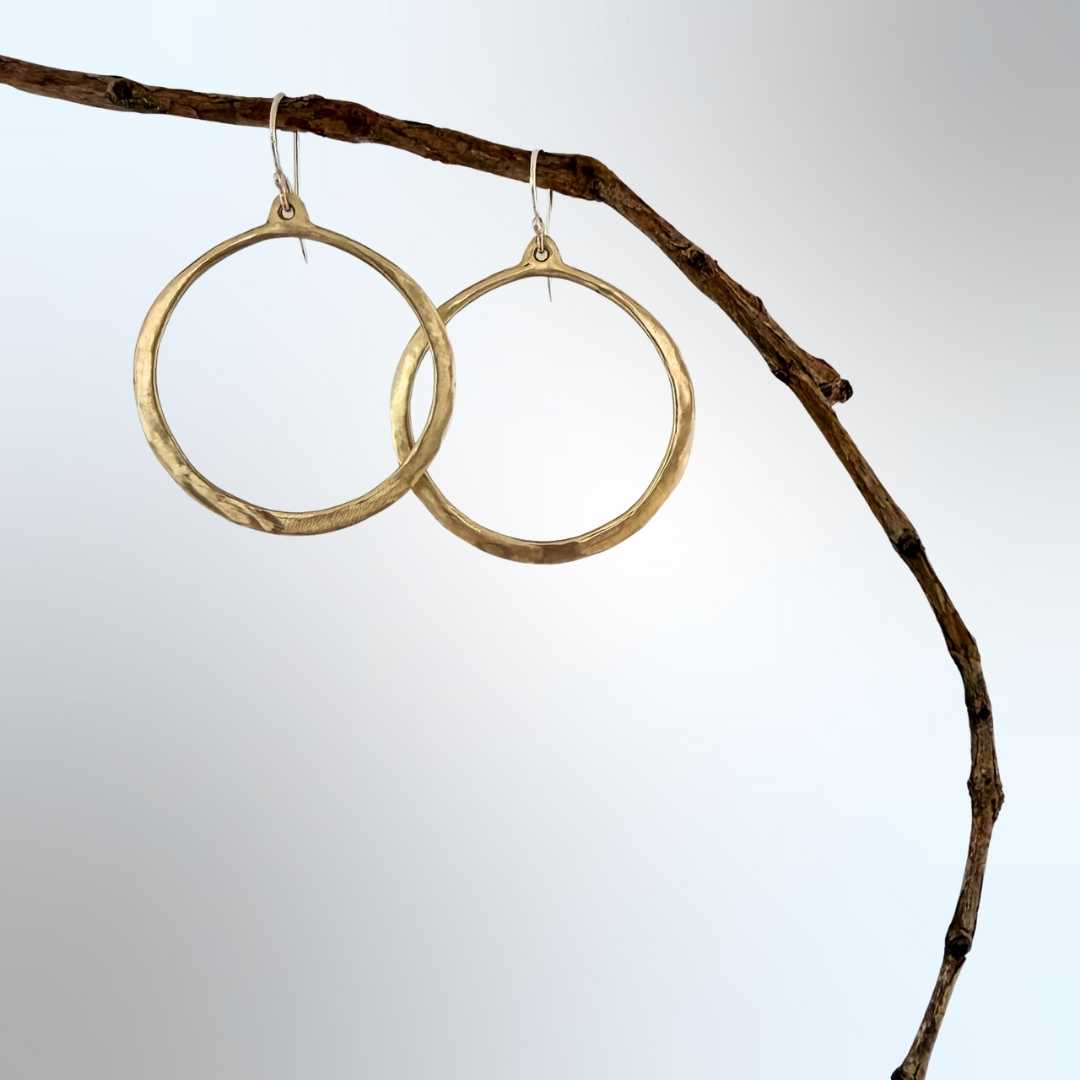 Lunar Eclipse hammered bronze hoop earrings with sterling silver hooks, hanging from a natural wood branch against a soft blue-grey background, hoops slightly overlapping with one sitting lower than the other.
