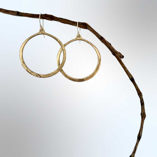 Lunar Eclipse hammered bronze hoop earrings with sterling silver hooks, hanging from a natural wood branch against a soft blue-grey background, hoops slightly overlapping with one sitting lower than the other.