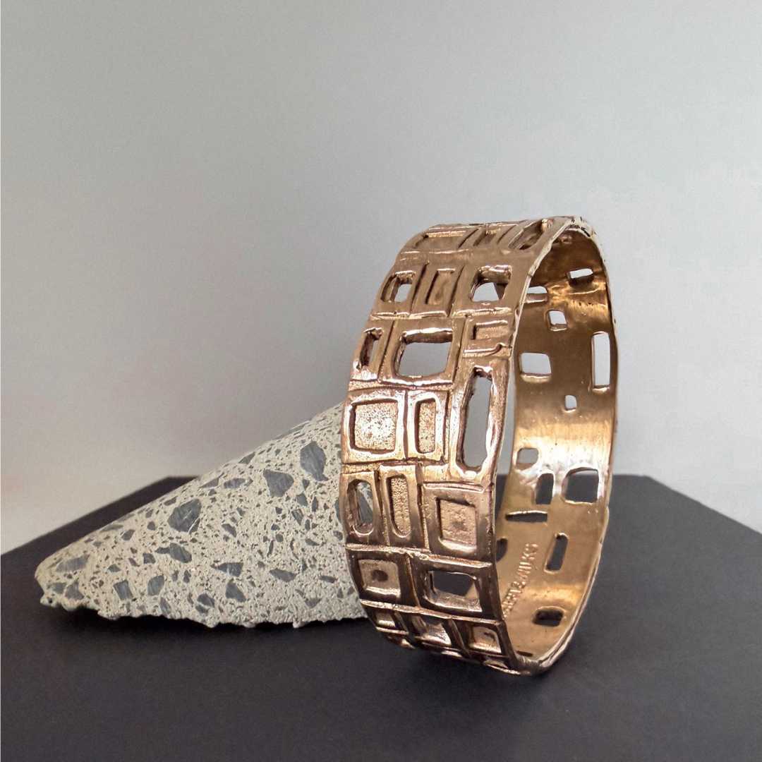 HerbertandWilks Modernist Bangle in warm, caramel-toned bronze, standing on its side to display the geometric cut-out design against a textured concrete wedge.