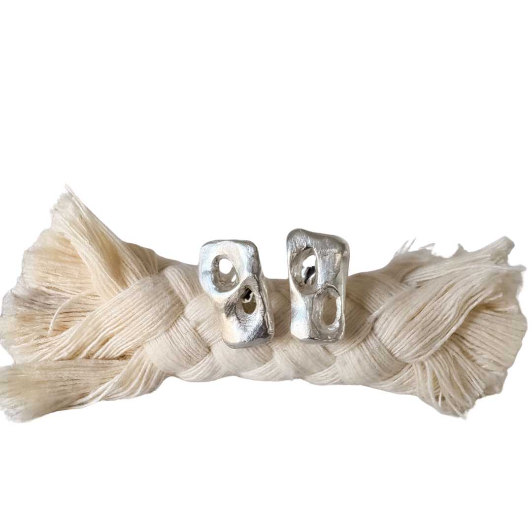 Sterling silver Perfora Rectangle Stud Earrings shown worn the opposite way, resting on a braided rope against a white background, emphasising their asymmetric design