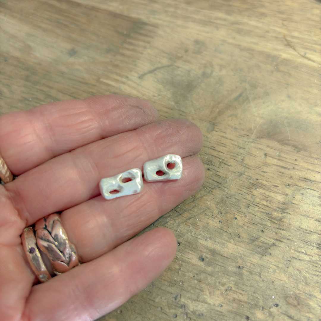 Sterling silver Perfora Rectangle Stud Earrings shown from a distance, lying face up on a hand against a wooden background