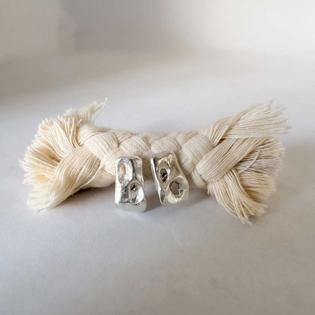 Sterling silver Perfora Rectangle Stud Earrings resting on a braided rope against a variegated grey background, showing sculptural pierced surface