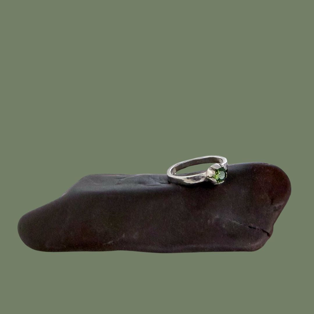 Handcrafted 925 sterling silver stacking ring featuring a round green peridot stone, displayed on a dark brown polished stone against a solid olive green background.