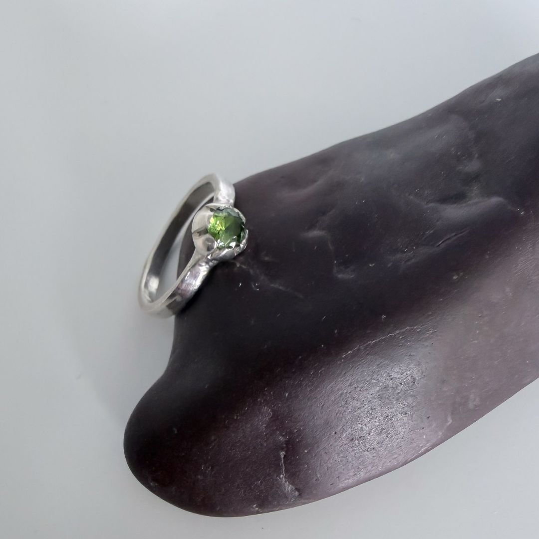 Sterling silver Peridot Bloom Stacking Ring featuring a 5mm round lime green peridot gemstone in a botanical claw setting, displayed on a dark brown polished stone against a light gray background.