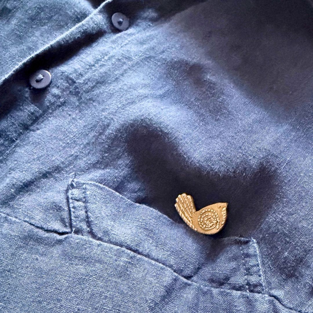 Bronze Pīwakawaka Fantail Pin worn on a dark blue linen shirt pocket
