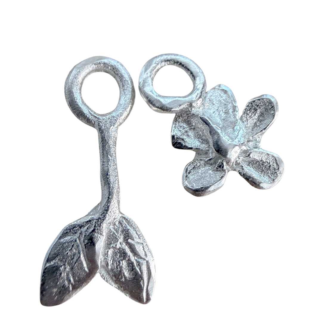 A detailed close-up of two sterling silver botanical earring charms—one plum blossom and one leafy stem—by HerbertandWilks. Handmade local Auckland jewellery displayed on a white background.