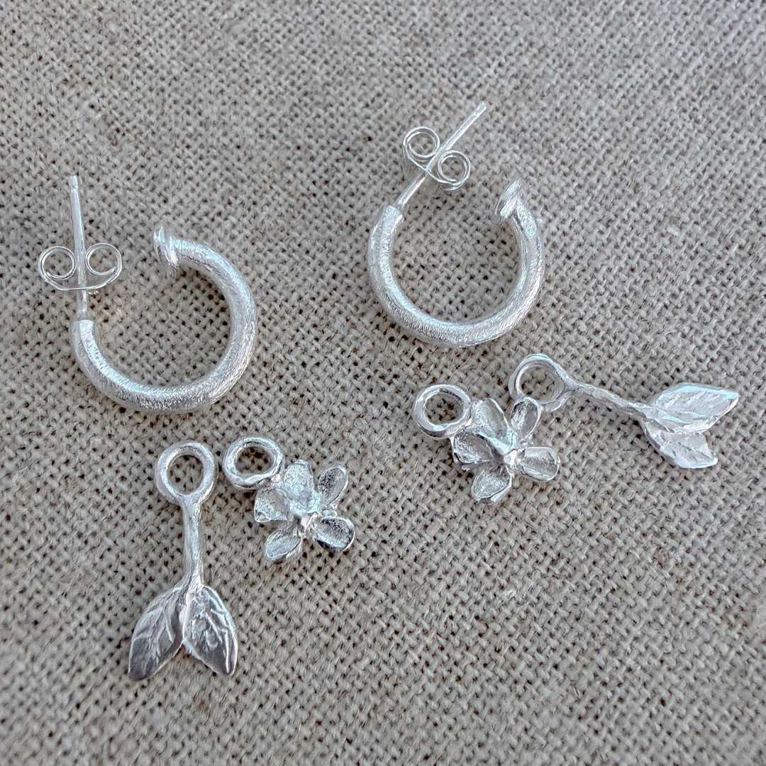 Handcrafted sterling silver Plum Blossom & Leaf earring full set bundle by HerbertandWilks Jewellery. This local Auckland jewellery piece includes textured silver hoops with two flower and two leaf charms, shown on a beige linen background.