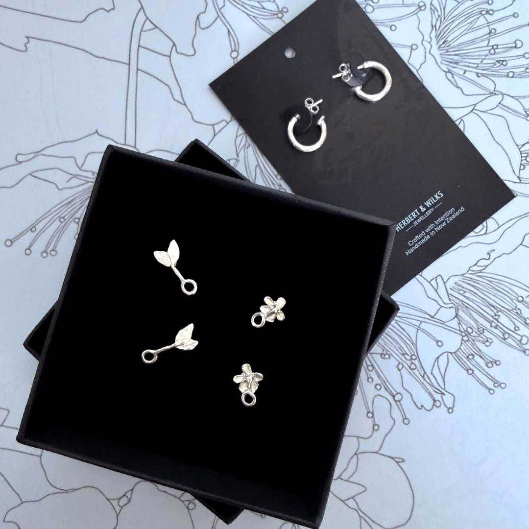 Handcrafted sterling silver Plum Blossom & Leaf earring full set by HerbertandWilks Jewellery, displayed in a black branded presentation box. This local Auckland jewellery set includes hoops and charms, shown against a light blue floral background.