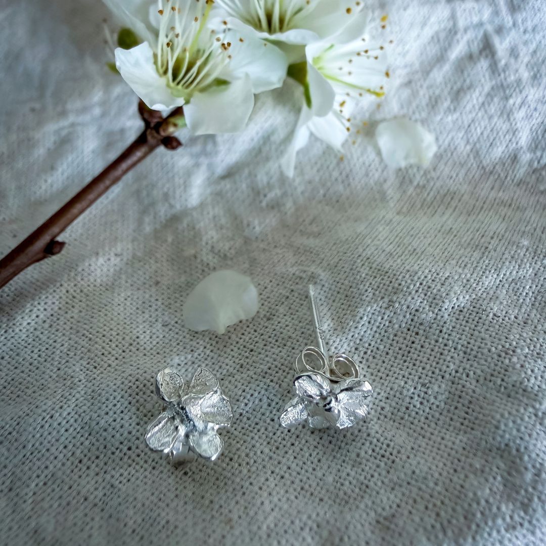 Sterling silver plum blossom stud earrings beside fresh white plum blossoms, with one earring showing its post and butterfly back.