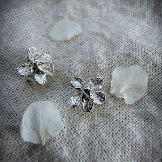 Sterling silver plum blossom stud earrings on natural linen with scattered white plum blossom petals.