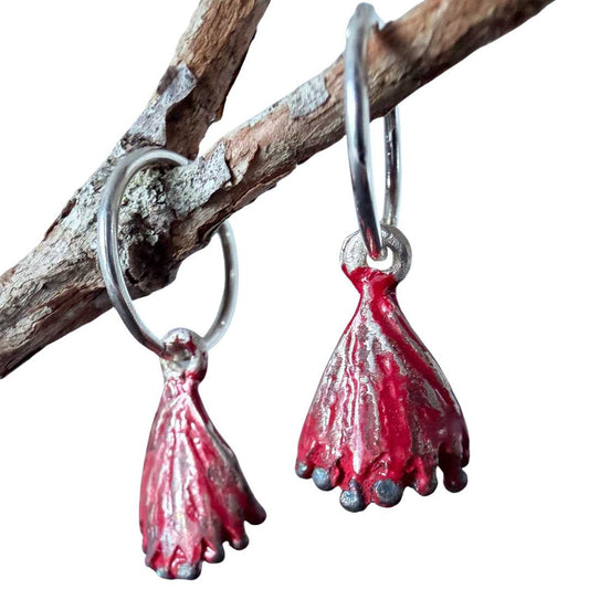 Macro detail of handmade Pohutukawa earrings hanging from a branch, showing the dappled red enamel finish and oxidised silver stamens.