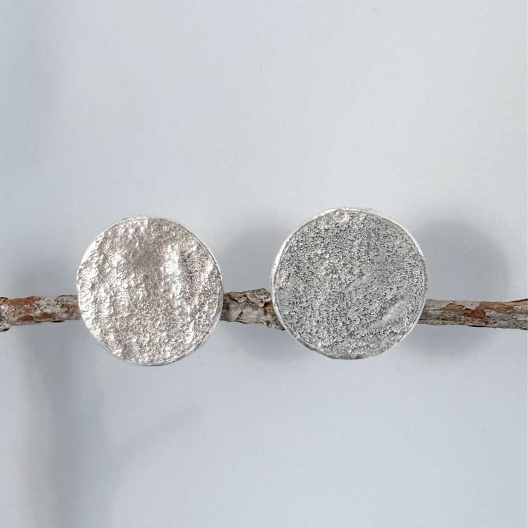 Minimalist view of the sterling silver Sequin Disc Stud Earrings resting on a straight horizontal branch against a pale grey background, with their distinct shapes silhouetted by soft shadows on the back wall.