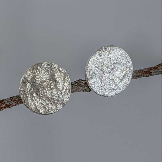Artistic shot of the sterling silver Sequin Disc Stud Earrings balanced at staggered levels on a gently upward-sloping branch against a dark grey background, emphasising the organic, light-diffusing texture.