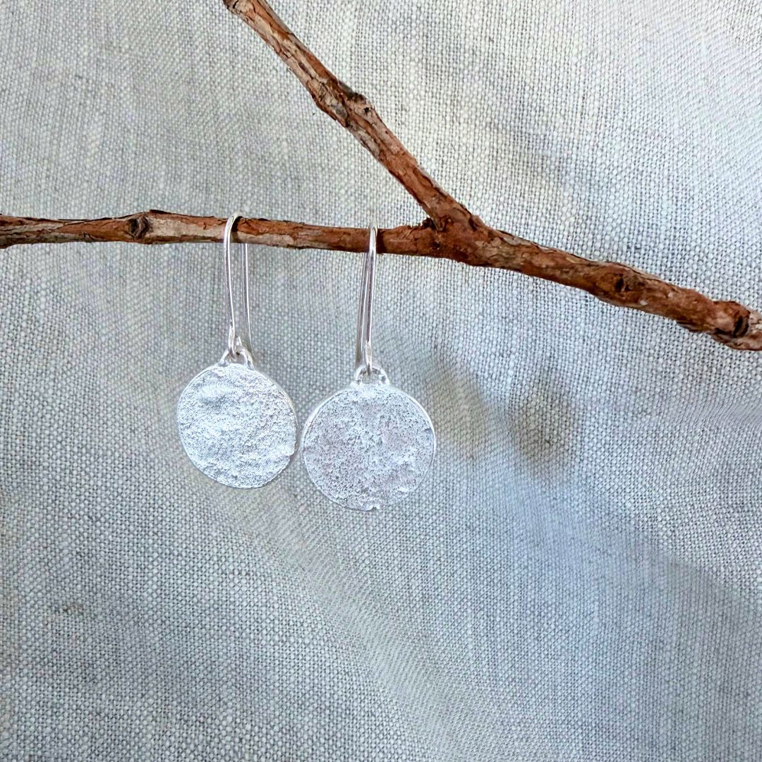 A pair of Sequin Swing sterling silver dangle earrings with textured circular discs hanging from simple silver hooks, displayed against a neutral linen background with a small brown branch across the top.