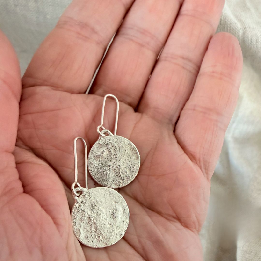 A pair of sequin swing earrings showing the signature textured silver discs with fishhook wires, resting on an open hand against a soft neutral fabric background.
