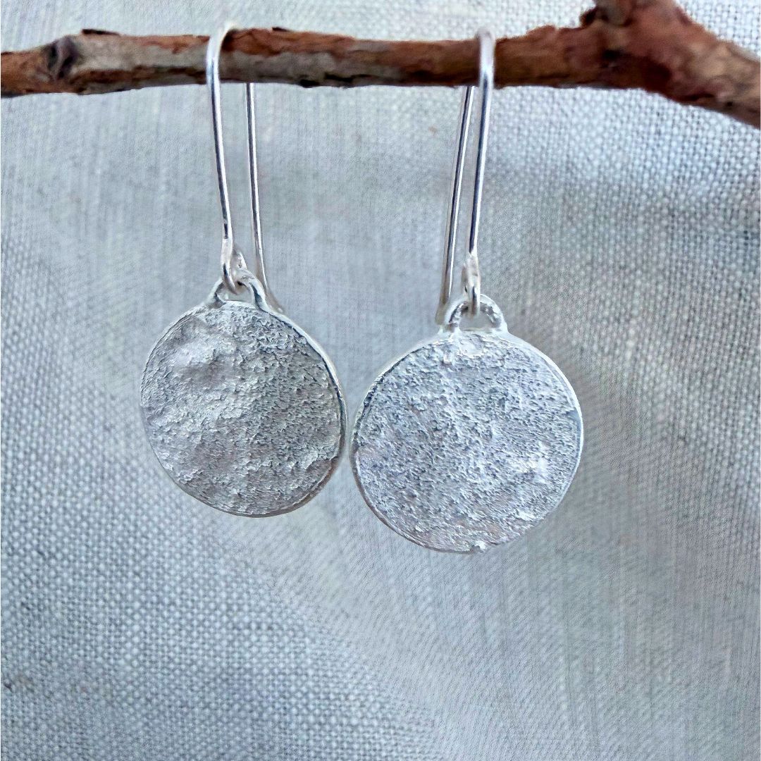A pair of textured sterling silver Sequin Swing Earrings - simple disc drop earrings hanging from simple silver fishhook wires, displayed against a neutral, light gray linen fabric backdrop with a rustic brown twig branch across the top.