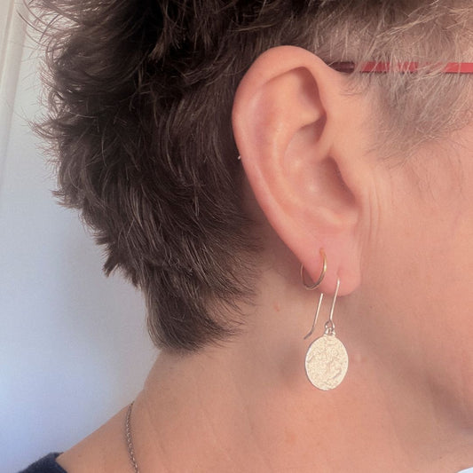 A close-up of a person's ear wearing a small gold hoop earring and the Sequin Swing earring with its signature textured circular disc, against a light background.