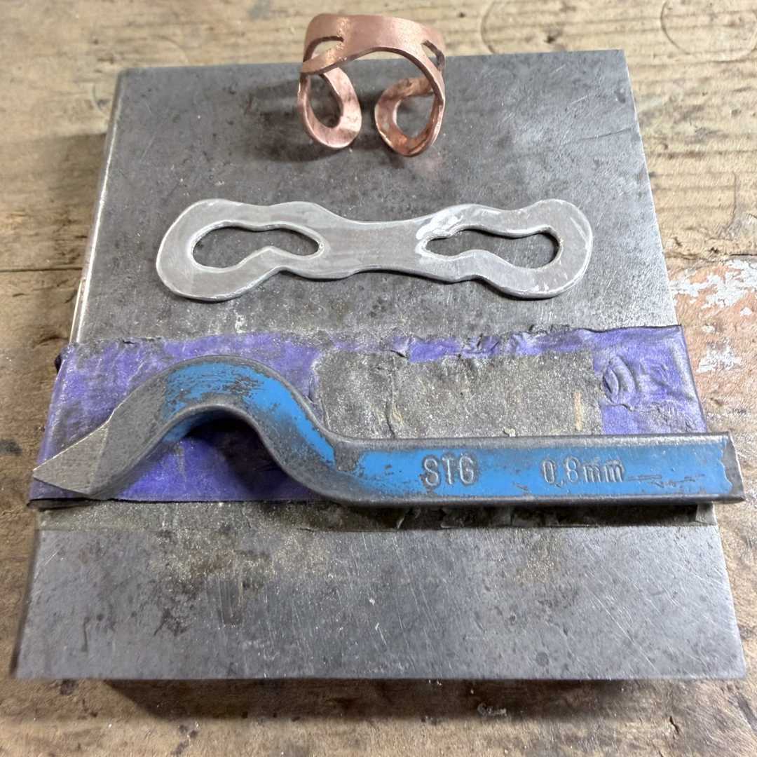 Sterling silver sheet about to be stamped with the “STG” hallmark, cut and textured while still flat lying on a steel block on a jewellery workbench - a sample copper ring sits alongside
