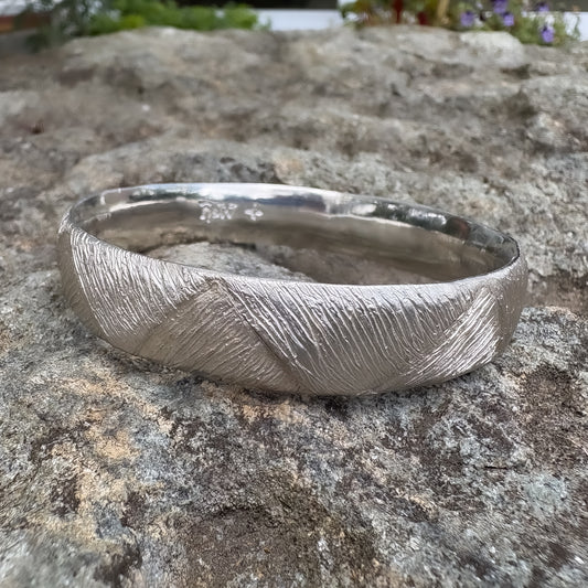 Strata Silver Bangle Edition of One resting on basalt rock with greenery in soft focus — showing the woven surface texture and engraved HerbertandWilks marks.