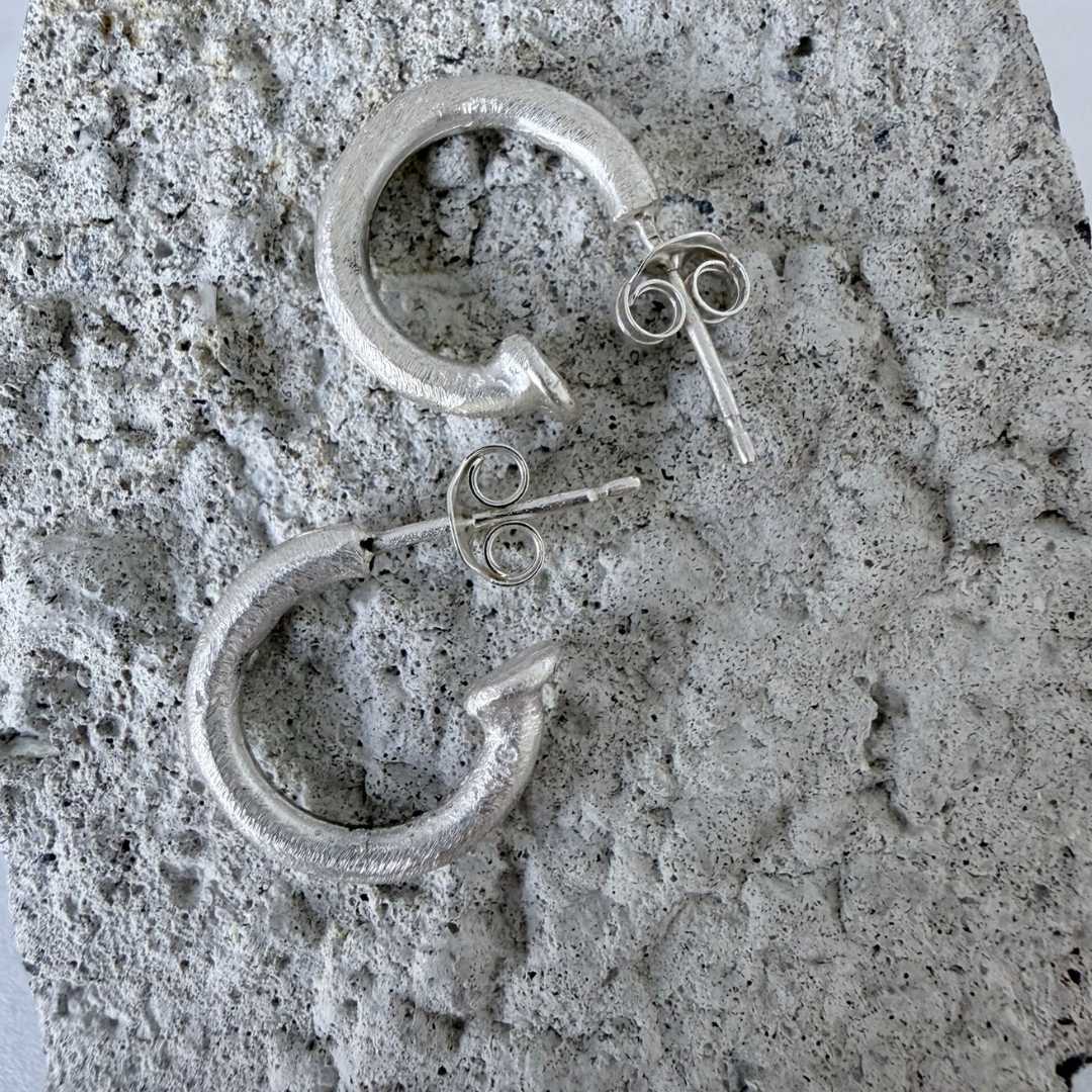 14mm textured sterling silver hoop earrings lying on textured concrete surface highlighting hand-finished texture