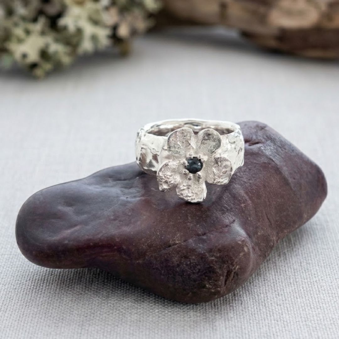 Front view of the Waiatarua Wide sterling silver flower ring on dark stone, with a deep blue sapphire at its centre.