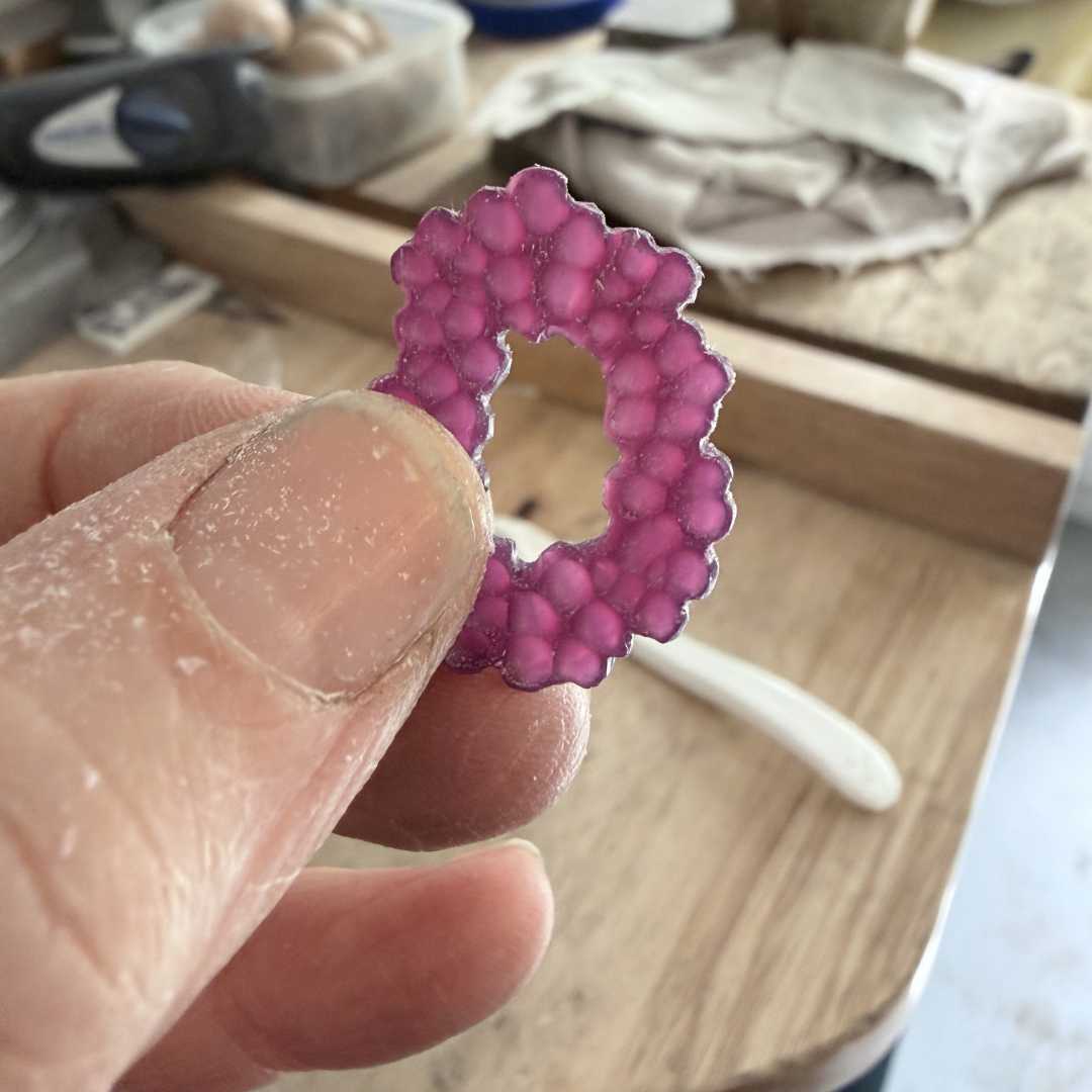 A close-up of a maker's fingers holding an organic, bubbly-textured purple wax model for a pendant or earring at a jeweller's bench.