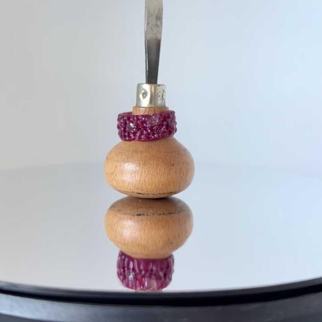 A textured purple wax ring model with embedded gemstones, resting on a wooden-handled jeweller's carving tool over a mirrored reflective surface.