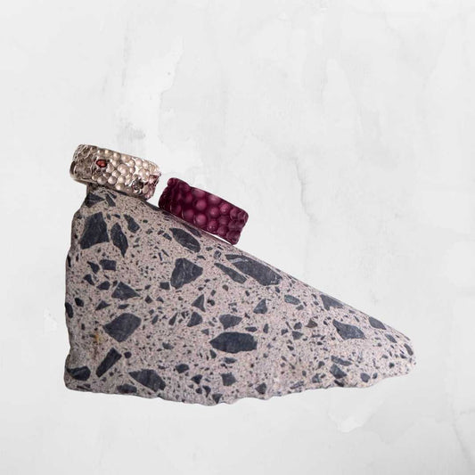 A comparison showing a purple carved wax ring model next to the finished textured sterling silver ring with a small red gemstone, displayed on a speckled terrazzo stone wedge.
