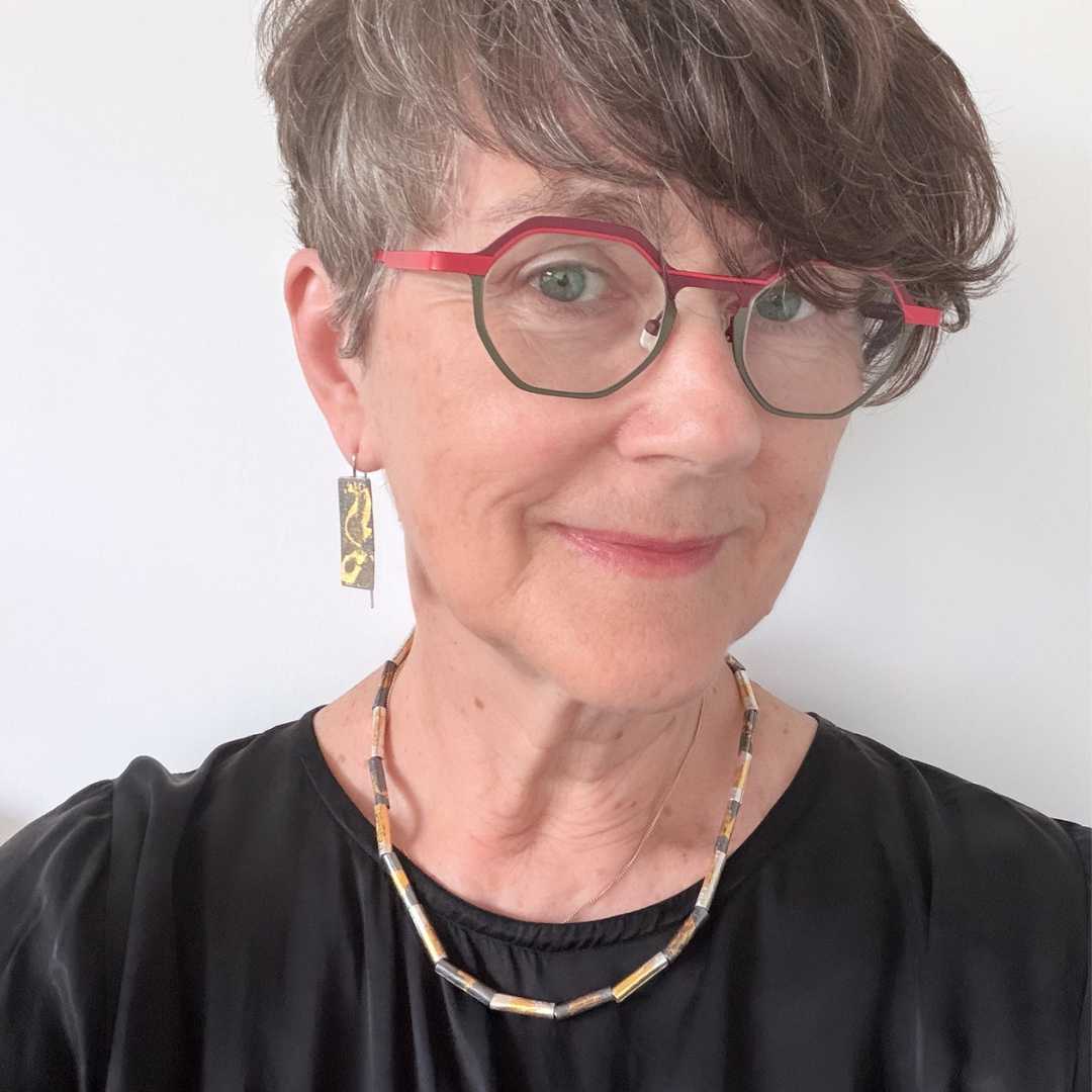 Woman with short hair wearing red and green glasses and a black dress, wearing wrapped fine silver and gold beaded necklace with matching gilded earrings