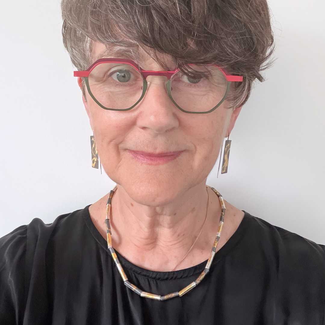 Woman with short hair wearing red and green glasses and a black dress, looking at camera, wearing wrapped fine silver and gold beaded necklace layered with a gold chain and matching gilded earrings