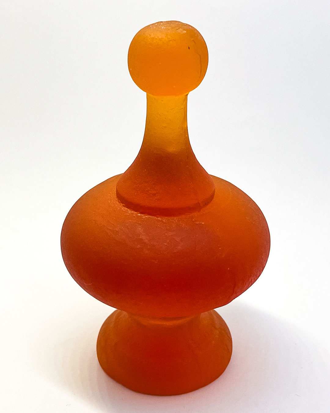 Close-up of the orange cast glass finial sculpture, showing its vibrant colour, textured surface, and sculptural form against a white background.
