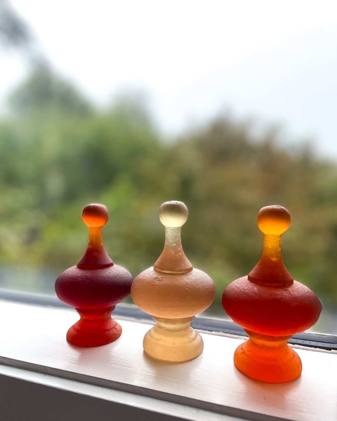 Rhubarb pink cast glass finial positioned between two dark orange finials on a window ledge, with natural light highlighting their colours and translucent forms.