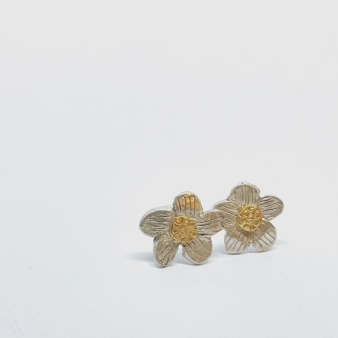 Daisy Stud Earrings propped up by their sterling silver posts, highlighting the recycled 18ct gold centres and handcrafted silver petals against a light grey surface.