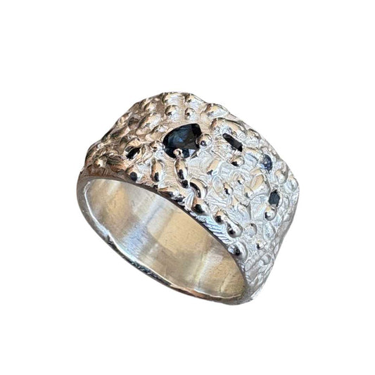 Sapphire Flow Wide Band Ring No. 23 photographed on a white background, shown from a top-down angled view. The image highlights the textured sterling silver band, the pear-shaped blue sapphire set into the surface, and a glimpse of the inside of the wide band.