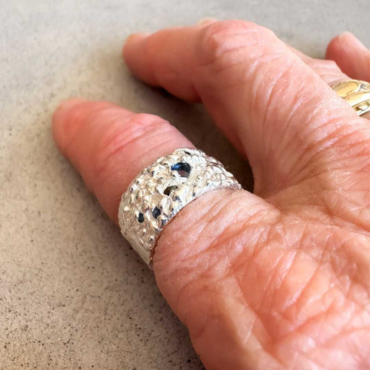 Flow Sapphire Wide Band Ring No. 23 worn on the hand against a neutral speckled concrete background, showing the textured sterling silver band and deep blue sapphire set into the surface.