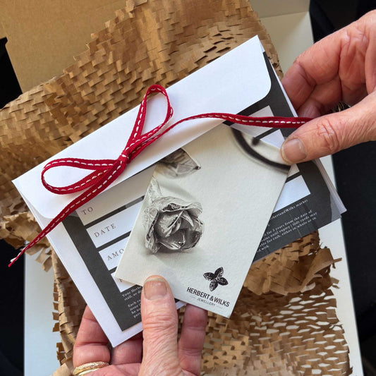Hands holding a HerbertandWilks Jewellery Gift Card with red-bow envelope in front of a post box — New Zealand gift idea