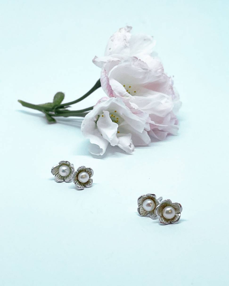 Two pairs of small flower Stud Earrings in Sterling Silver set with a white freshwater pearl, sitting in front of some blossom flowers