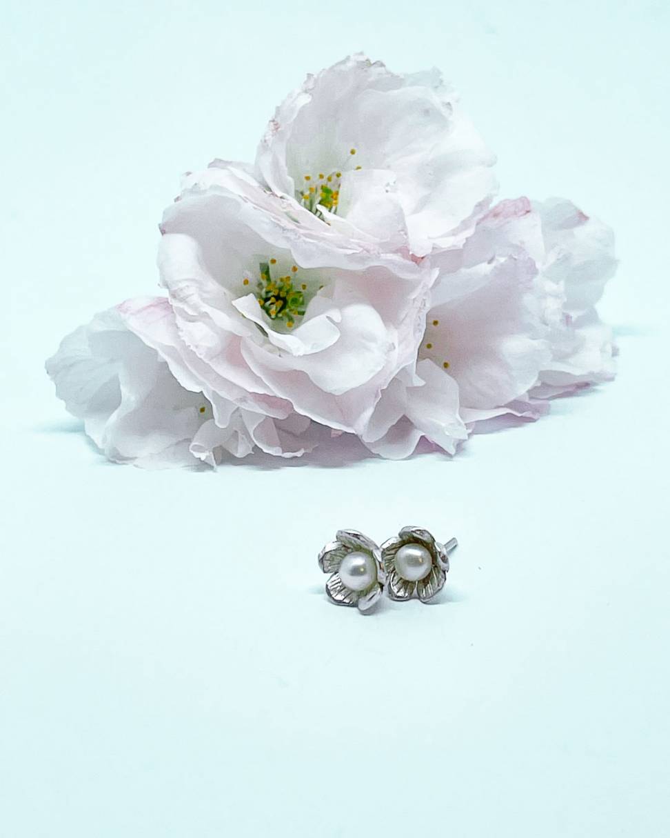 A pair of small flower Stud Earrings in Sterling Silver set with a white freshwater pearl, sitting in front of some blossom flowers