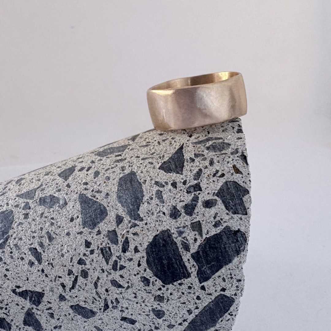 Wide band organic ring in 9ct yellow gold with satin finish, displayed on concrete core sample edge showing textured stone chips against medium grey background