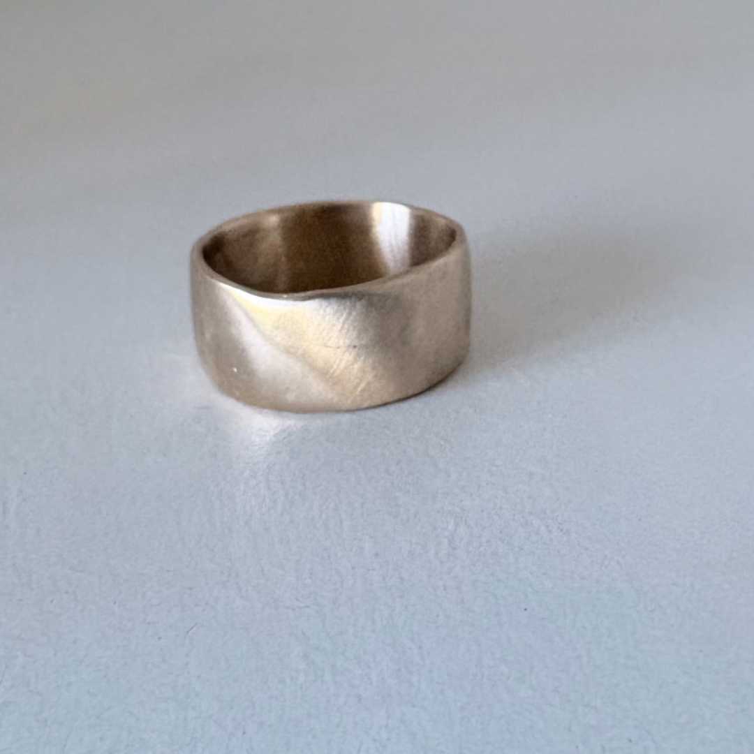 Wide band organic ring in 9ct yellow gold laying flat on light grey background, showing front surface and interior of tapered band with satin finish