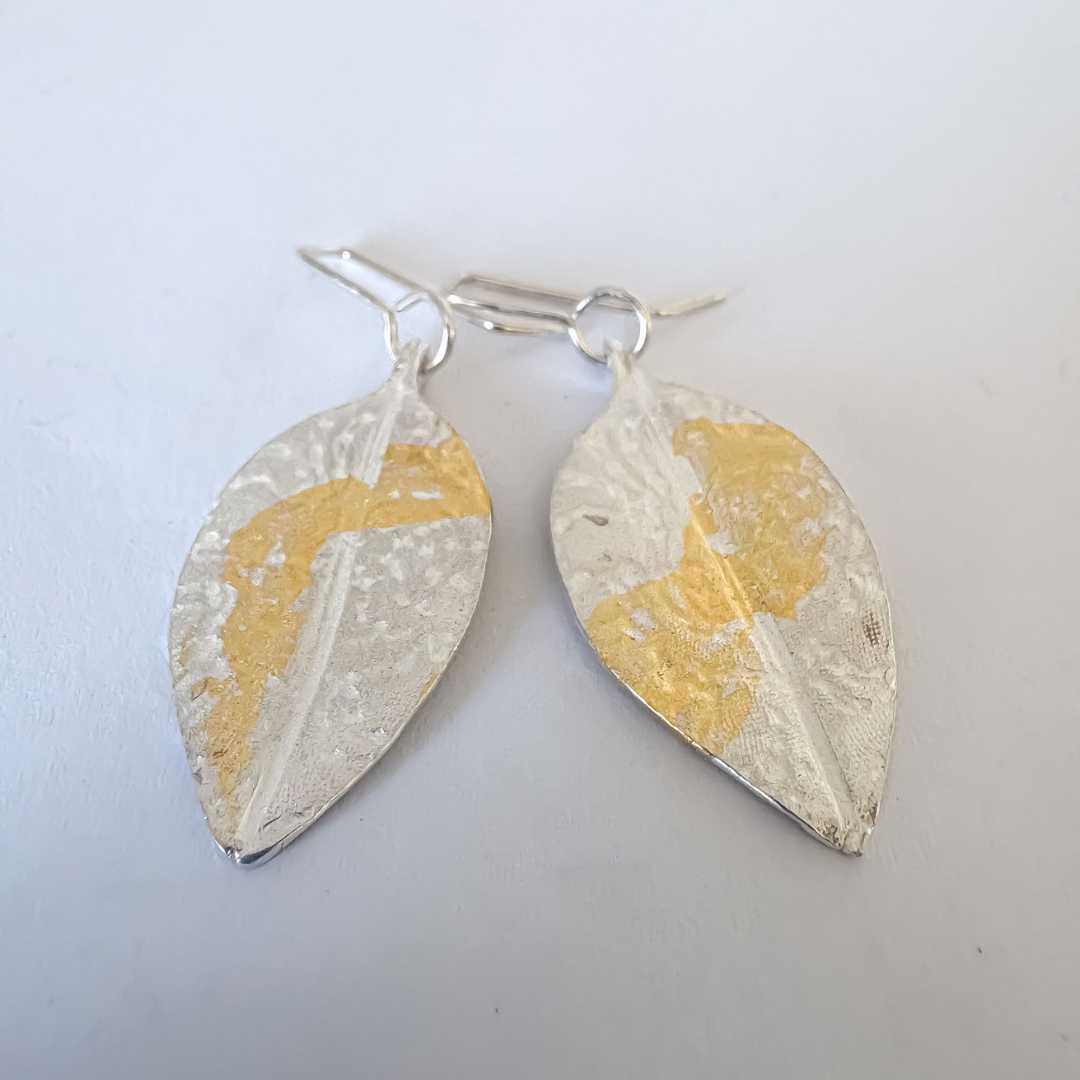 Top-angle view of Pohutukawa Lace Leaf Earrings showing fine silver texture and gold leaf detail on a light-grey background.