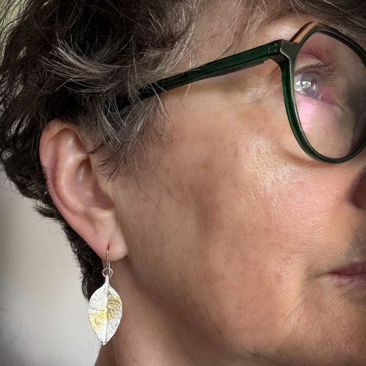 Woman wearing Pohutukawa Lace Leaf Earrings – fine silver and gold leaf dangles – short brown hair and glasses, Herbert and Wilks Jewellery.