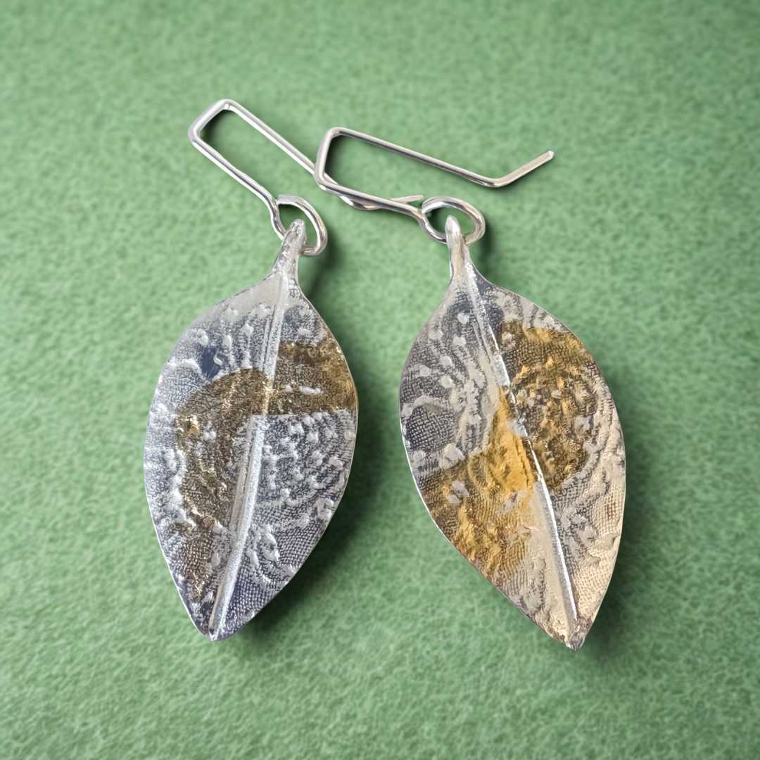 Pohutukawa Lace Leaf Earrings in fine silver and 24 ct gold leaf lying on a sage-green textured surface.