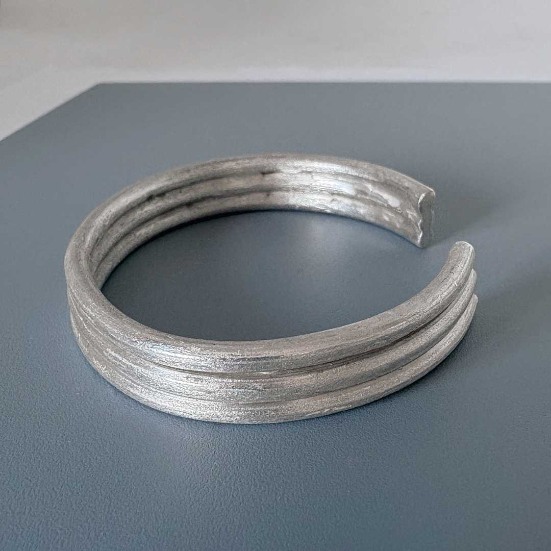 Side view of the Three Line Cuff in sterling silver resting on a steel-blue board with a light grey border, showing the profile, curve, and thickness of the design
